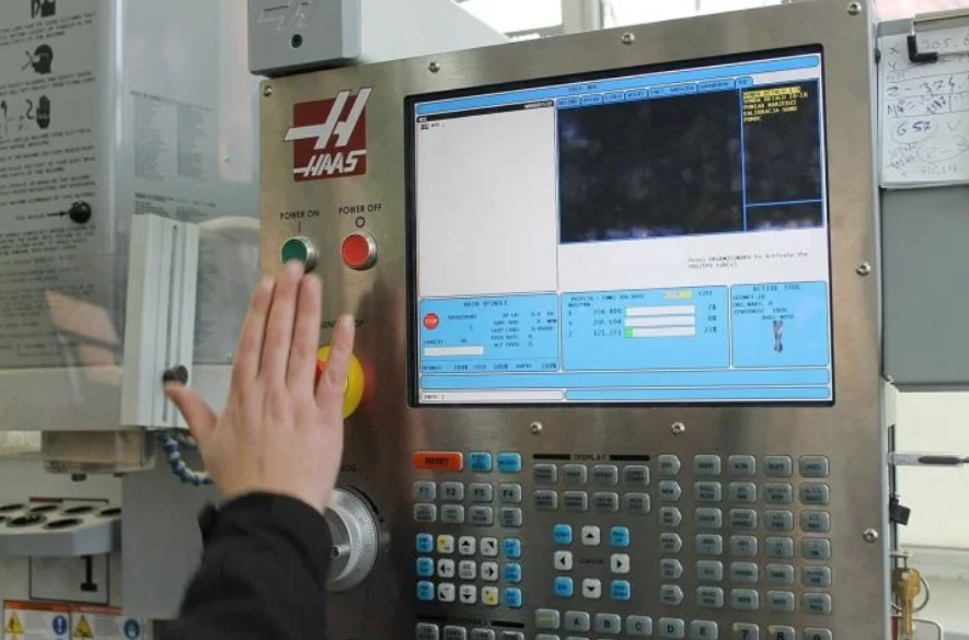 A closeup of a CNC machine control panel showing a program on the screen and a person about to press the green button 
