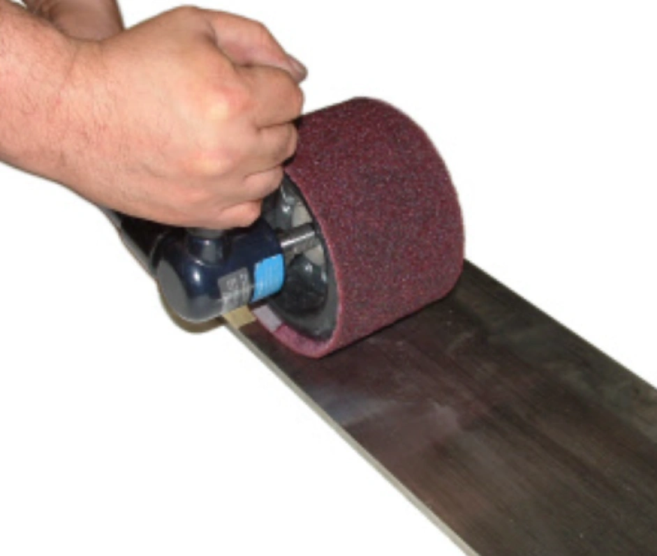Hand holding a belt sander, grinding a metal surface to refine grain structure in the graining process